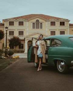A woman in a beige overcoat standing next to a green vintage car in front of a large building